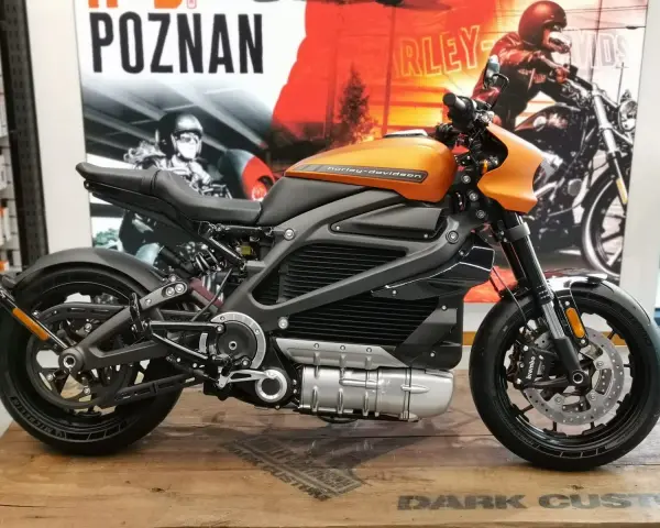 Pomarańczowy harley elektryczny z czarnymi akcentami, prezentowany na drewnianej podłodze, z plakatem Harley-Davidson w tle.