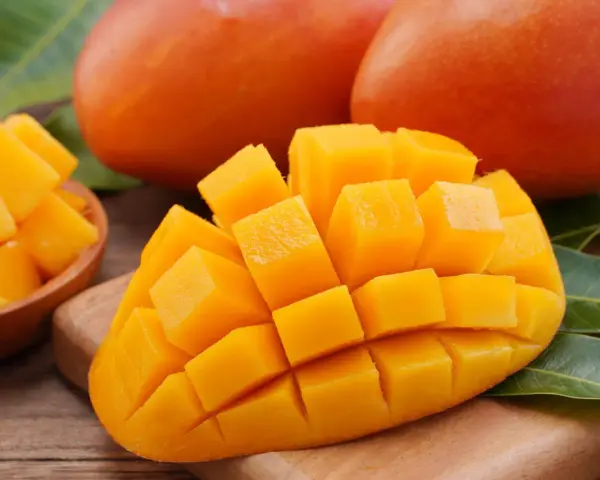 Dojrzałe mango, pokrojone w kostkę, prezentuje swoje soczyste, pomarańczowe wnętrze. Obok leżą całe owoce i liście.