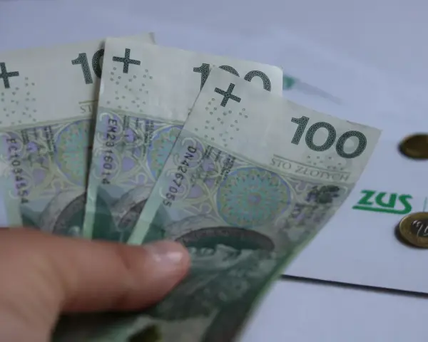 Trzy stuzłotowe banknoty w dłoni, obok dokument ZUS i monety. Czy to zapowiedź 30 krotności ZUS 2025?