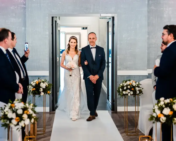 Panna młoda w sukni ślubnej idzie z ojcem do ołtarza, obok bukiety kwiatów.