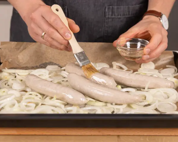 Bratwurst im Backofen: Zwiebelringe auf Backpapier, darauf vier rohe Bratwürste. Mit einem Pinsel wird Öl aufgetragen.