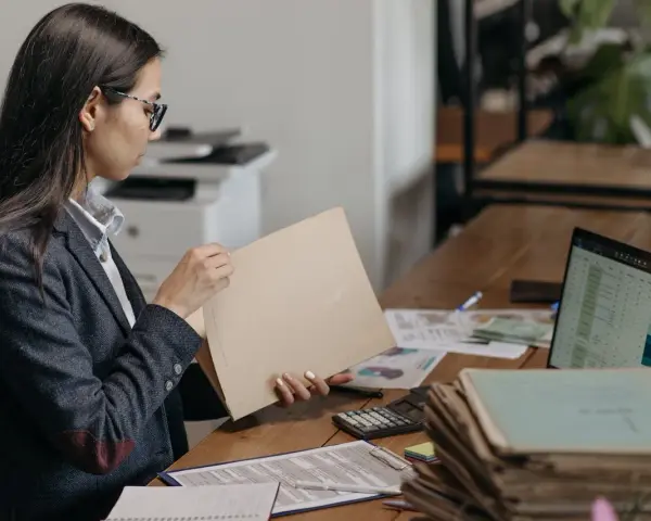 Kobieta w okularach przegląda dokumenty, szukając tytułu prawnego do lokalu. Na biurku leżą też laptop, kalkulator i sterty papierów.