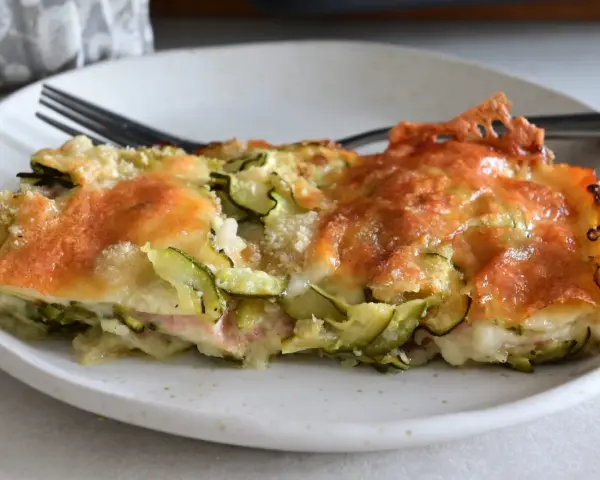 Una fetta di sformato di zucchine dorato e filante, con formaggio fuso e zucchine a rondelle, servito su un piatto bianco.