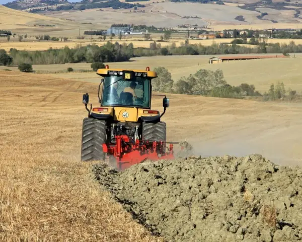 Trattore giallo in azione per l'aratura del terreno, sollevando terra e polvere in un paesaggio rurale collinare.