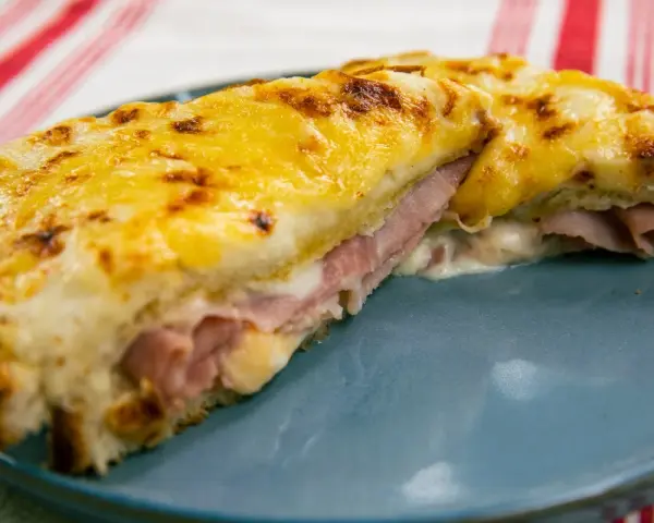 Croque Monsieur gratinado con queso derretido y jamón, servido en plato azul.