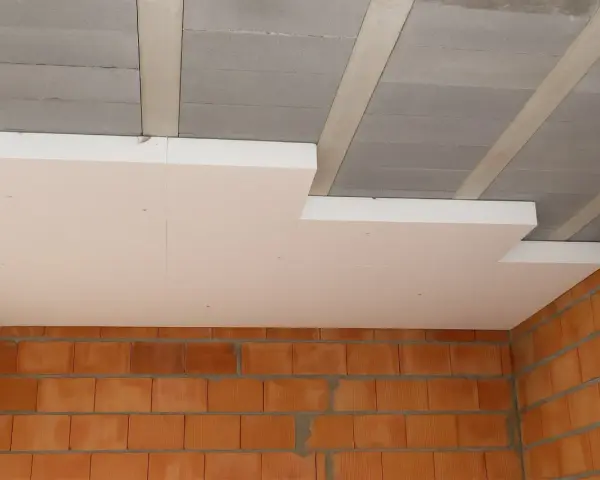 Ocieplenie stropu styropianem w trakcie budowy. Widoczne płyty styropianowe pod sufitem, betonowe belki stropowe i ściany z cegły.