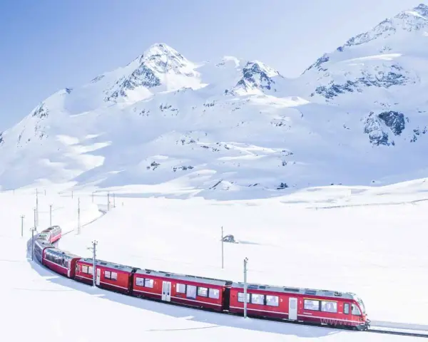 A bright red St. Moritz train winds through a pristine, snow-covered mountain landscape under a clear blue sky.