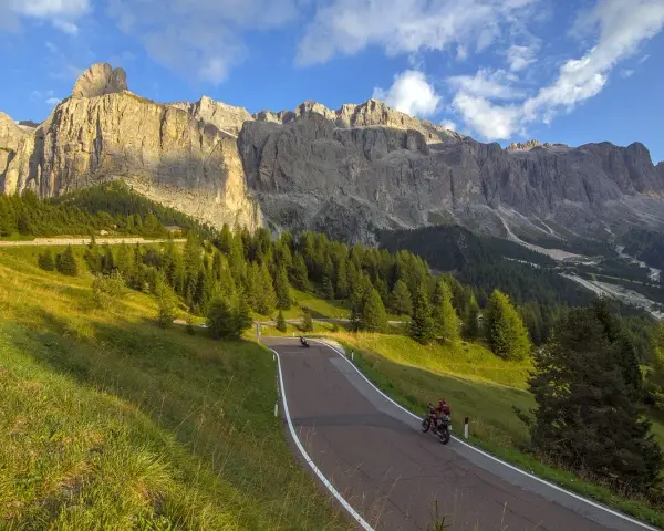Motociclisti sfrecciano su una strada tortuosa verso il Passo Furcia, circondati da verdi pendii e imponenti montagne rocciose sotto un cielo azzurro.
