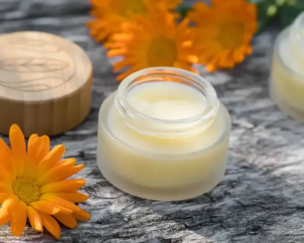 Ringelblumensalbe gegen Falten: Natürliche Pflege für glatte Haut. Kleine Gläser mit Salbe und leuchtend orangefarbene Ringelblumenblüten auf Holz.
