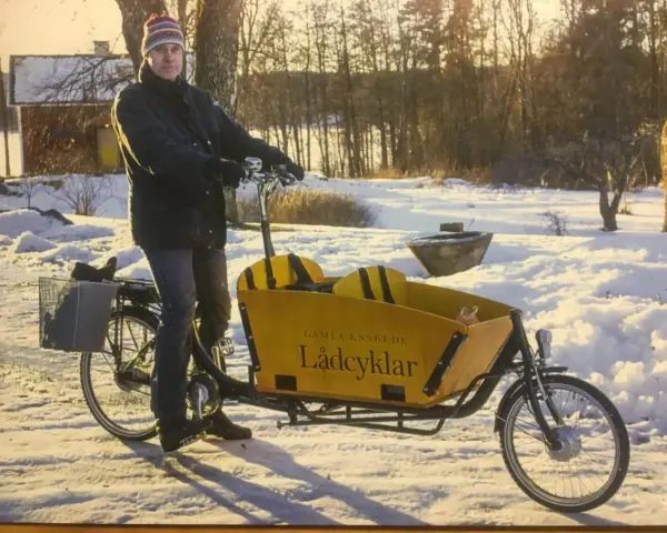 Man med mössa och jacka cyklar på vintern med en gul lastcykel i snöig miljö.
