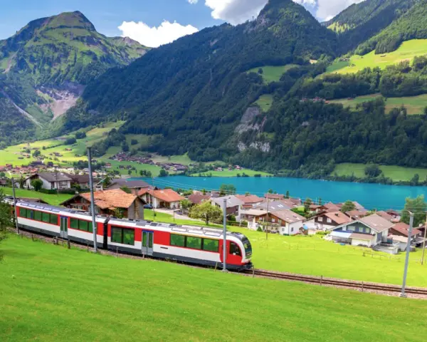 A red and white train travels through a picturesque Swiss village nestled beside a turquoise lake, with lush green mountains in the background. This is the scenic route from Lucerne to Lugano.