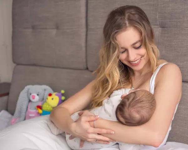 Uśmiechnięta mama karmi piersią dziecko. W tle pluszowe zabawki. Nakładki na pierś do karmienia mogą pomóc w tym intymnym momencie.