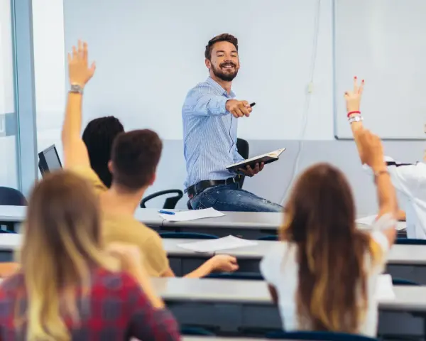 Uśmiechnięty wykładowca z książką w ręku wskazuje na studentów, którzy podnoszą ręce. Czy przed lecz stawiamy przecinek?