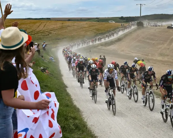 Spectators cheer as a peloton races on a dusty road during the Tour de France watch. Cyclists in colorful jerseys kick up dust in a dramatic scene.