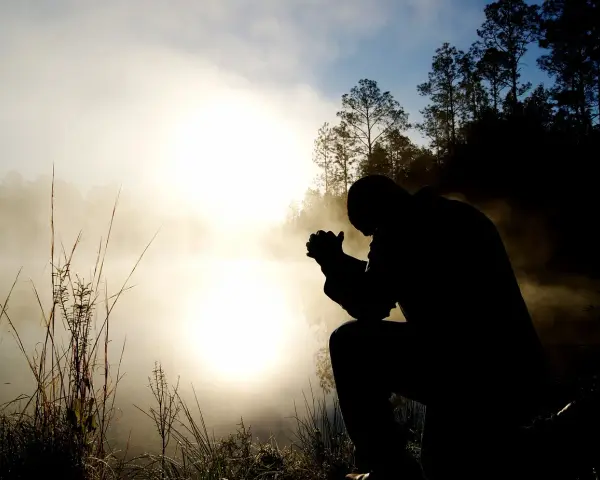 Silhouette eines Mannes, der im Morgennebel kniet und betet. Ein Bild, das christliche Glaubensbekenntnisse widerspiegelt.