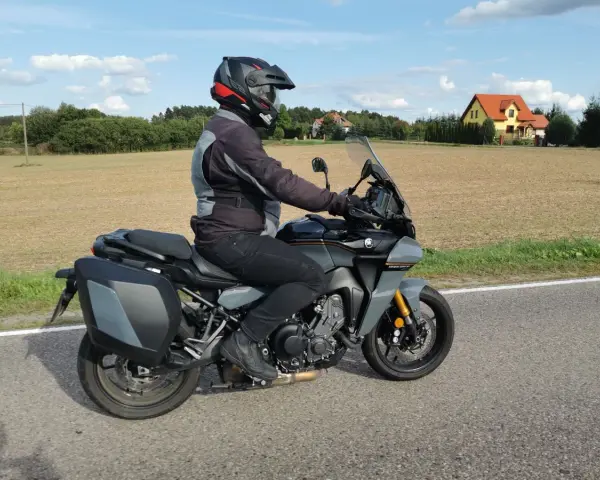 Motocyklista na szarym motocyklu Yamaha Tracer 900, z kuframi, w kasku, jedzie drogą. W tle pola i domy. Yamaha Tracer 900 dane techniczne.