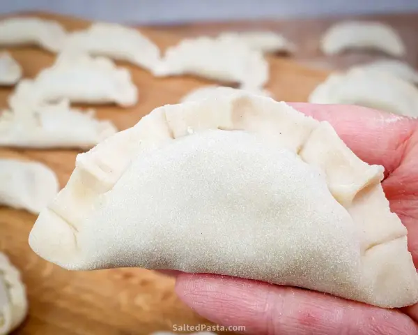 Dłoń trzyma surowe ciasto na pierogi, z innymi pierogami w tle.