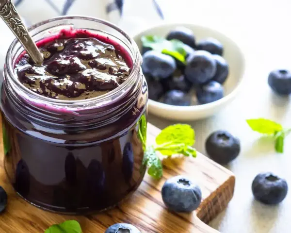 Vaso di marmellata di mirtilli fatto in casa, con mirtilli freschi e foglie di menta su un tagliere di legno.
