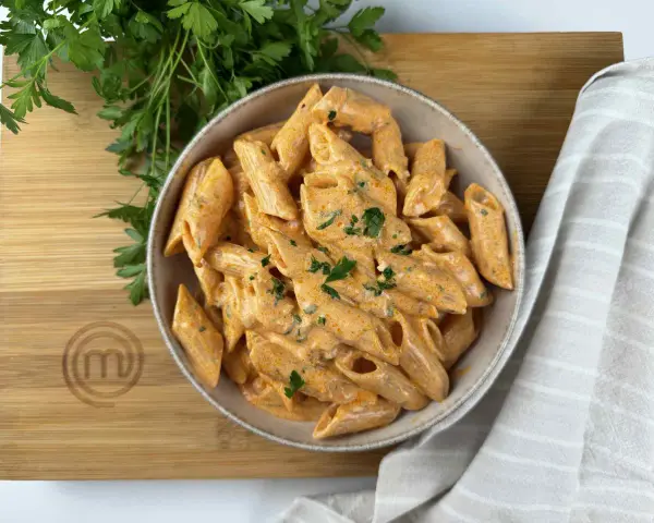 Penne alla vodka cremose e invitanti, guarnite con prezzemolo fresco, servite in una ciotola su un tagliere di legno con il logo MasterChef.