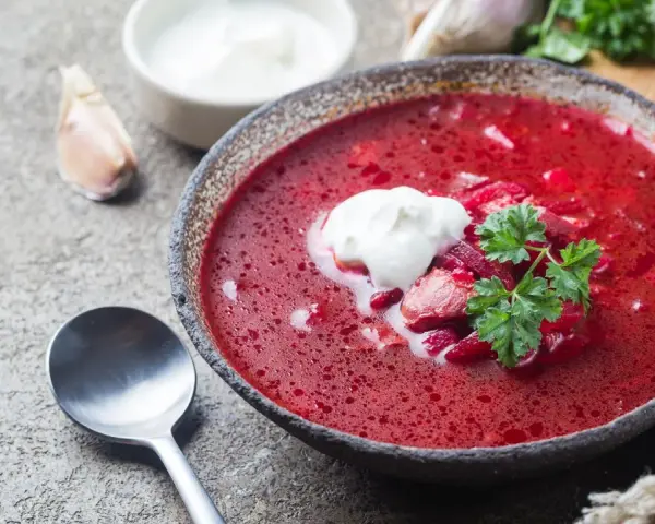Aromaticzny barszcz czerwony domowy z łyżką śmietany i natką pietruszki, obok czosnek i chleb.