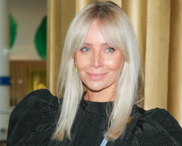 Blondynka z niebieskimi oczami i uroczą grzywką na boki, uśmiecha się lekko.