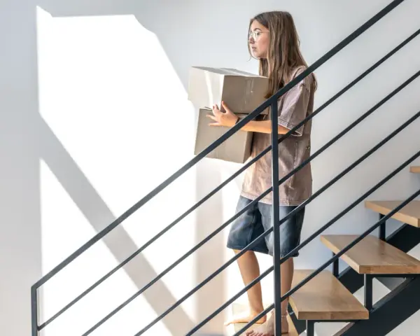 Nowa klatka schodowa w domu jednorodzinnym inspiracje: dziewczyna z kartonami schodzi po nowoczesnych schodach z drewnianymi stopniami i metalową balustradą.