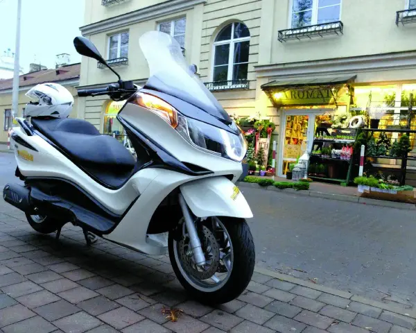 Biały skuter Piaggio X10 125, z kaskiem na siedzeniu, zaparkowany przed kwiaciarnią. Dane techniczne tego modelu są imponujące.
