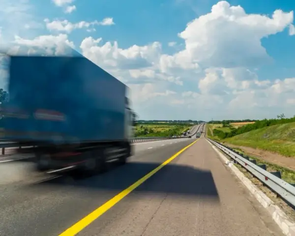 Niebieska ciężarówka w ruchu na autostradzie pod błękitnym niebem z chmurami.