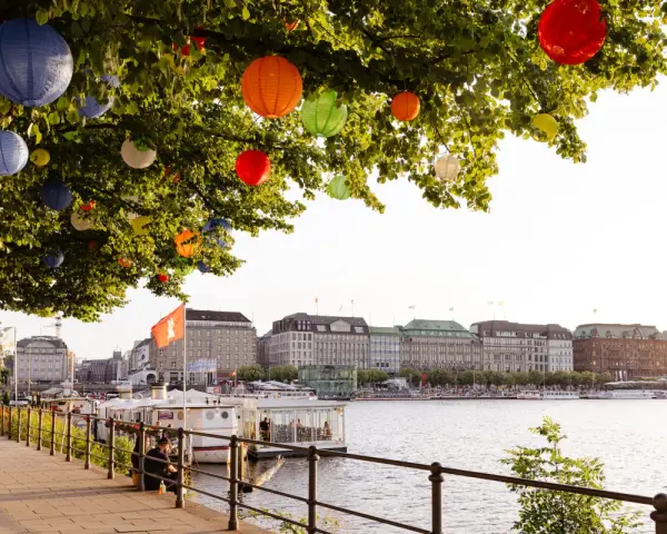 Bunte Lampions schmücken einen Baum am Wasser. Im Hintergrund die elegante Skyline von Hamburg mit Gebäuden und Booten.
