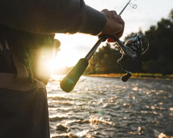Wędkarz trzyma wędkę z kołowrotkiem nad wodą. Słońce zachodzi, tworząc refleksy na falach. Zastanawiasz się, jaka plecionka do feedera będzie najlepsza?