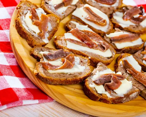 Na drewnianej desce ułożone są kromki chleba z masłem i anchois. To pyszne, proste przekąski.