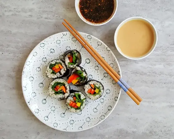 Sześć wegetariańskich rolek sushi z kolorowymi warzywami, przygotowanych z użyciem glonu do sushi, leży na talerzu obok sosów i pałeczek.