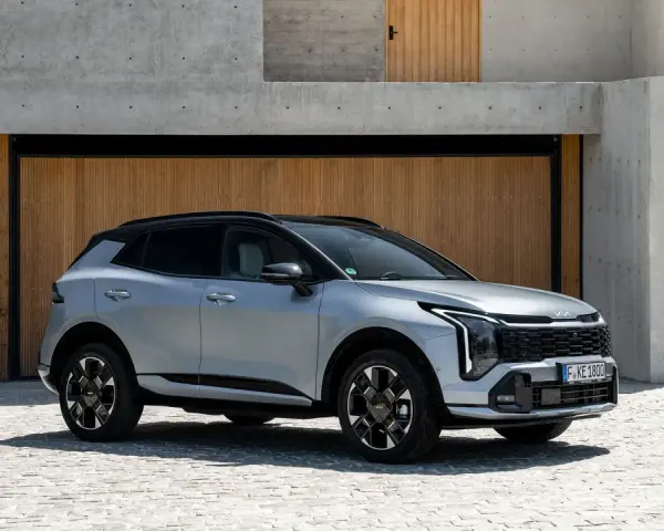 Elegancki, srebrny Kia Sportage Hybrid na tle nowoczesnej architektury z drewnianymi drzwiami i betonowymi schodami.