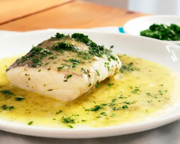 Un filete de merluza en salsa verde, con perejil picado, servido en un plato blanco.