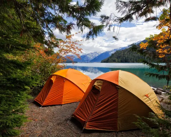 Två orangea tält står uppställda vid en sjö, omgivna av höstfärgad skog och berg. En perfekt plats för kamlunge camping.