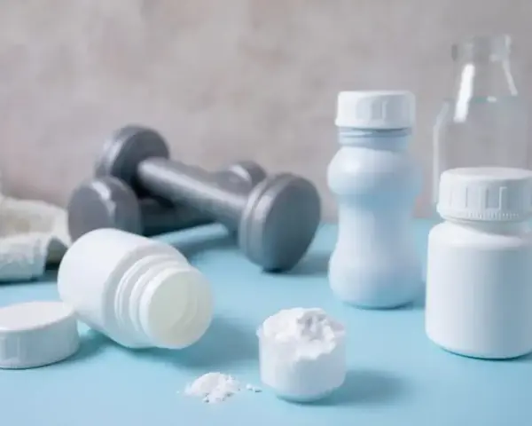 Białe pojemniki, miarka z proszkiem i hantle sugerują suplementację. Proteiny co to? To budulec mięśni, ważny dla sportowców.