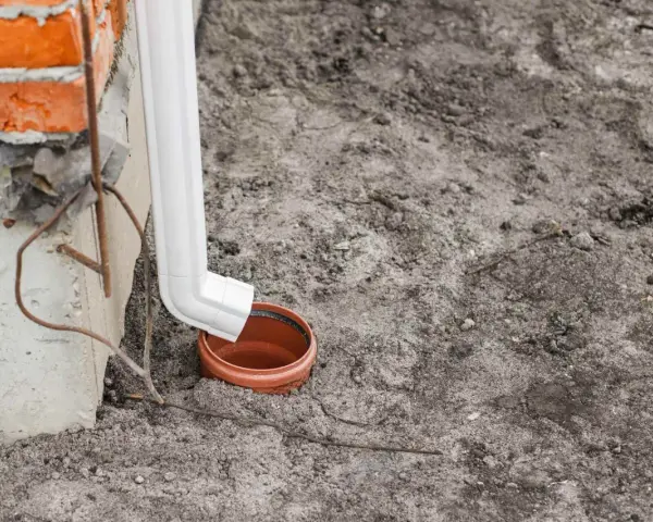 Biała rura odprowadza wodę z rynien do brązowej studzienki. Budynek z cegły w tle.
