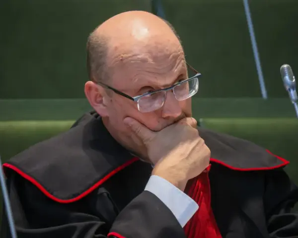 Sędzia w todze z czerwoną lamówką, w okularach, z ręką na brodzie, zamyślony.