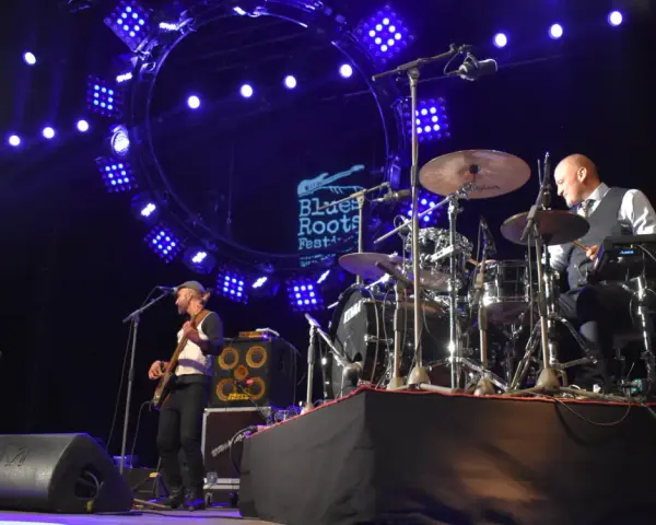 Un groupe joue du blues musique sous un grand cercle lumineux. Un bassiste à gauche, un batteur à droite.