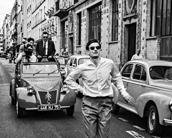 Un uomo con sigaretta in bocca, occhiali da sole e camicia a righe, in strada con auto d'epoca, evoca lo spirito de **la nouvelle vague**.