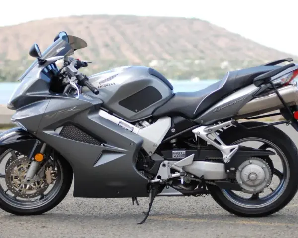Szary motocykl Honda VFR 800 z widokiem na wodę i wzgórze w tle.