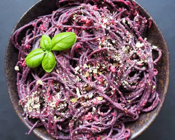 Fioletowy makaron spaghetti z buraczanym pesto, posypany orzechami i udekorowany listkiem bazylii.