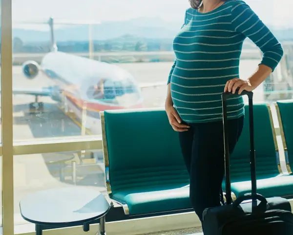 Ciężarna kobieta z walizką czeka na lotnisku. W tle widać samolot i pasażerskie fotele. Latanie samolotem w ciąży to wyzwanie, ale podróż dobiega końca.