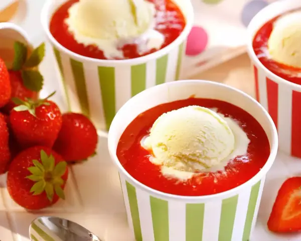 Vanilleeis mit Erdbeeren und Sahne in bunten Bechern. Frische Erdbeeren liegen daneben.