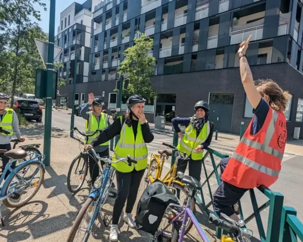 Des adultes souriants, casqués et en gilets fluo, prêts à apprendre à faire du vélo adulte. Ils sont avec leurs vélos, devant un immeuble moderne.