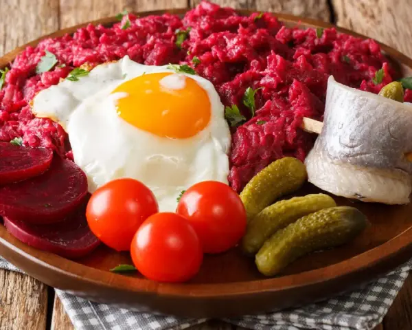 Herzhaftes Labskaus essen: Rote Bete, Spiegelei, Matjes, Cornichons und Tomaten auf einem Holzteller.