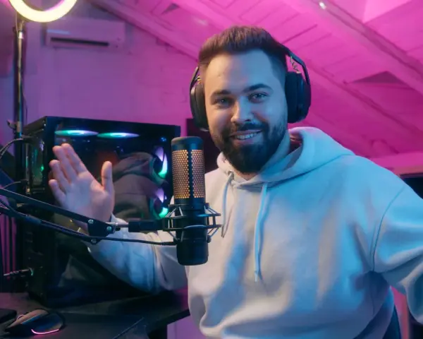 Uśmiechnięty streamer w słuchawkach, przy mikrofonie, tłumaczy, streaming co to jest. W tle komputer i różowe oświetlenie.