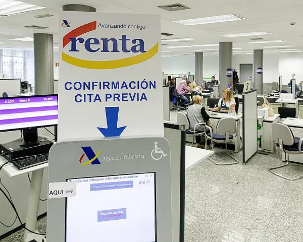 Oficina de la Agencia Tributaria con gente esperando. Un cartel indica "Confirmación cita previa" y una pantalla táctil para gestionar citas, resolviendo la duda de cuanto tarda Hacienda en cobrar la renta.