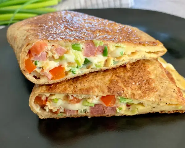 Keto tortilla z jajkiem, warzywami i wędliną, pokrojona na pół, prezentuje apetyczny przekrój.