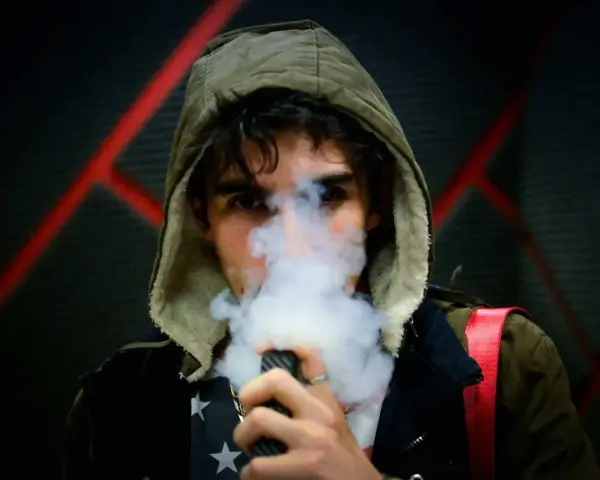 Joven con capucha exhala vapor de un vapeador, con un diseño de estrellas en su camiseta. Los nicokits son su secreto.